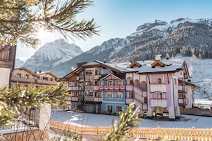 Hotel de varios pisos con balcones, suelo cubierto de nieve y montañas al fondo.