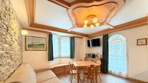 A cozy room with a stone wall, a wooden ceiling, a dining table with chairs, a sofa, and a TV mounted on the wall.