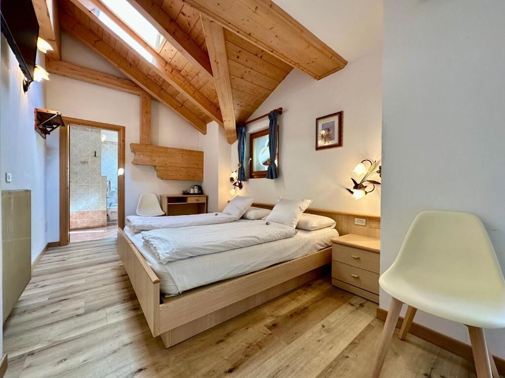 Un dormitorio con techo de madera, una cama, mesitas de noche, una silla y una puerta que da a otra habitación.