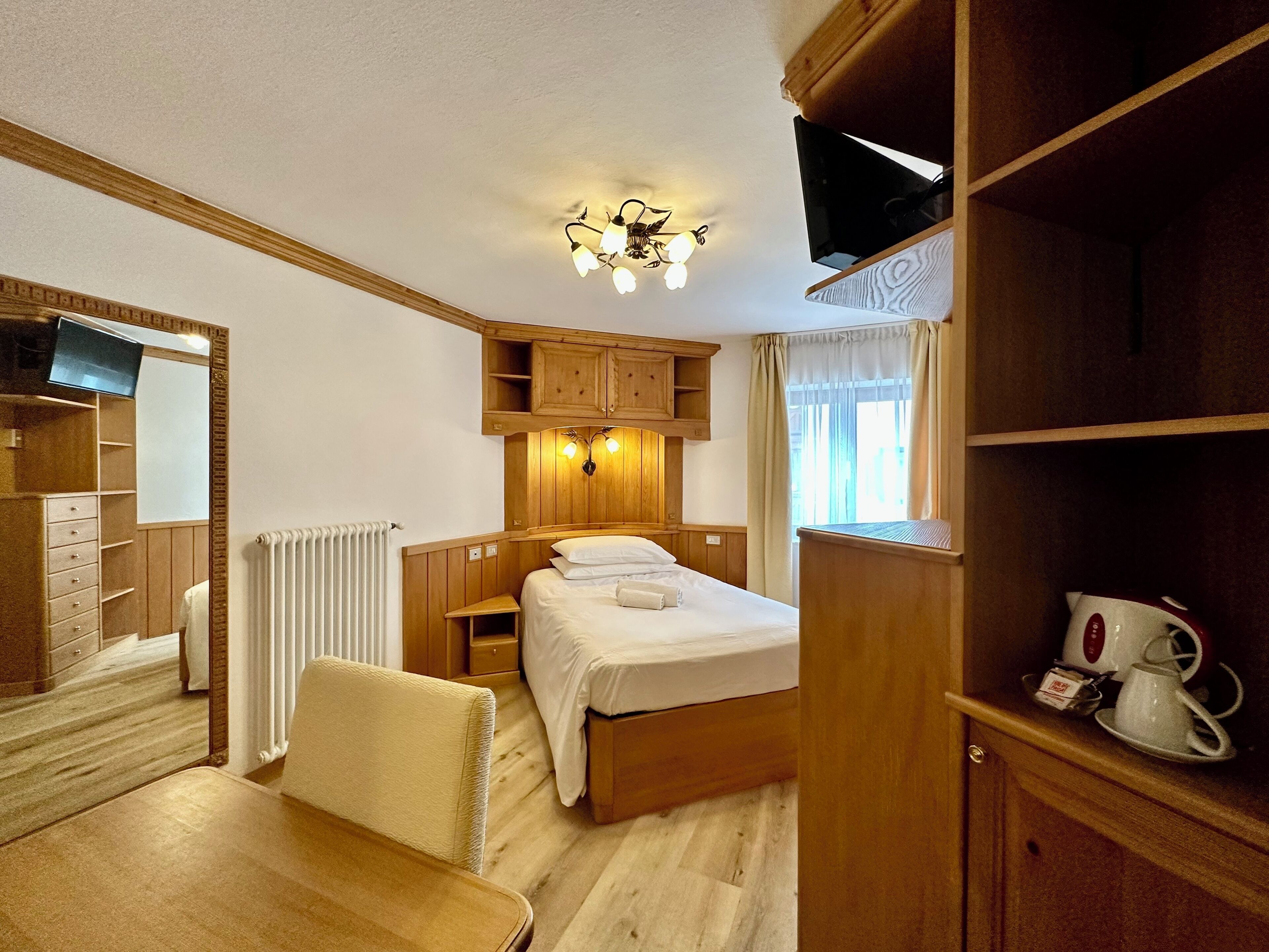 A hotel room with a bed, a wooden wardrobe, a desk with a chair, a television, and a radiator.