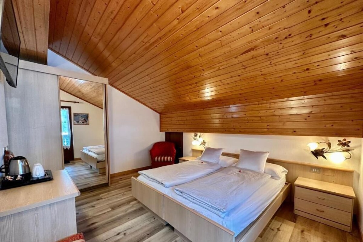 Un dormitorio con techo de madera, una cama con ropa blanca, una mesita de noche, una silla y un buró con una lámpara.