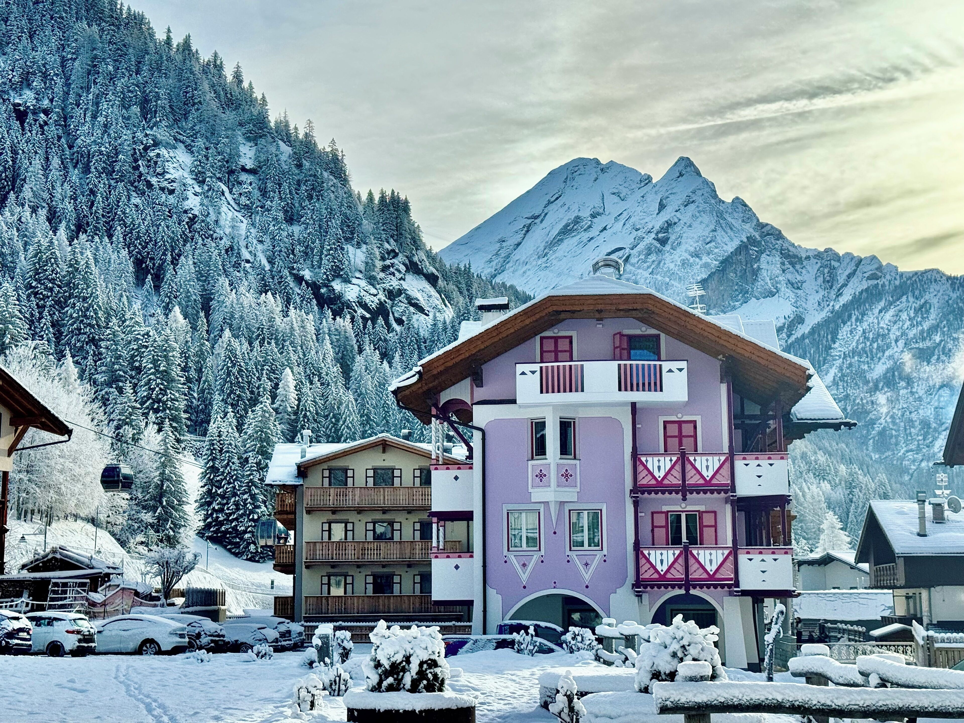 Un edificio de varios pisos con balcones, un telesilla al fondo y árboles cubiertos de nieve.