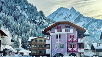 Un edificio de varios pisos con balcones, un telesilla al fondo y árboles cubiertos de nieve.
