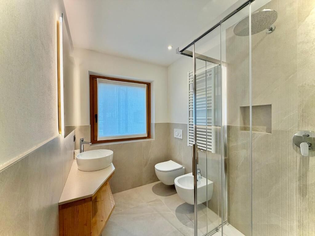 A modern bathroom with a glass shower enclosure, a large window, and a wooden vanity.
