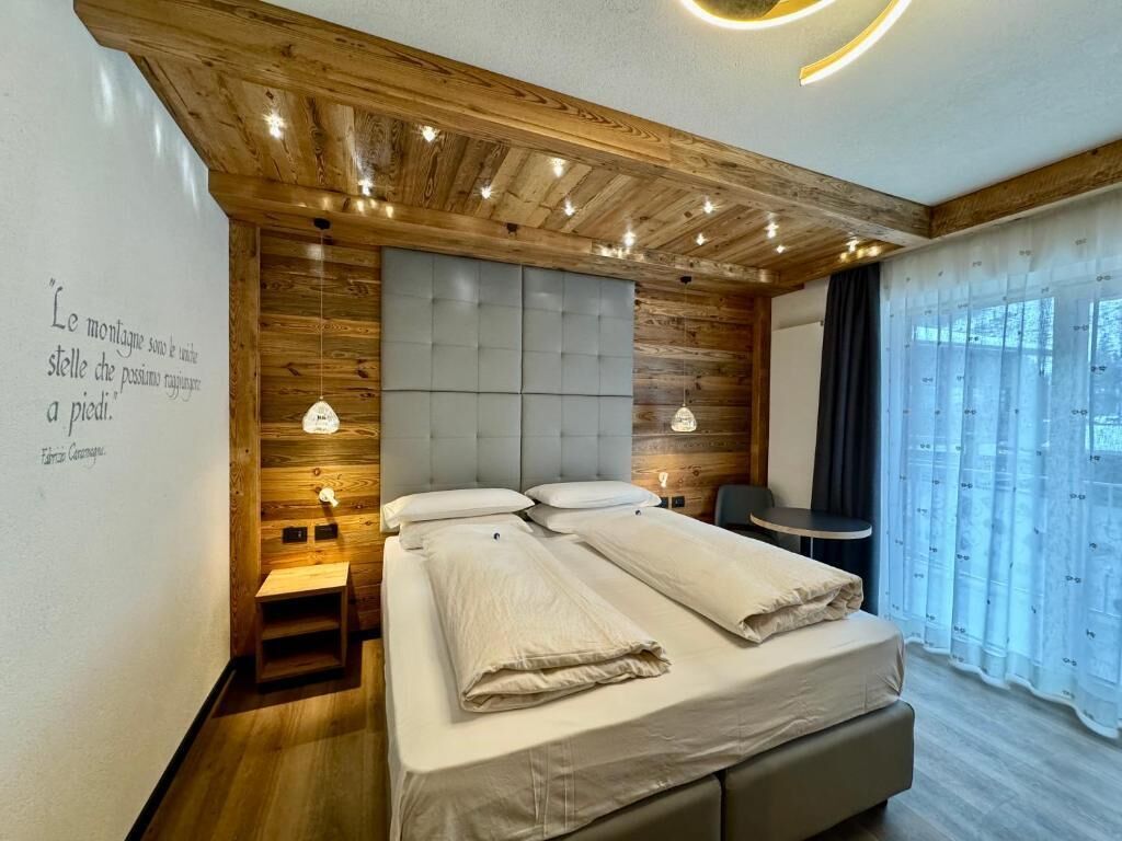 Un dormitorio con techo de madera, cabecera capitonada, cama con ropa de cama blanca, mesita de noche y ventana con cortinas.