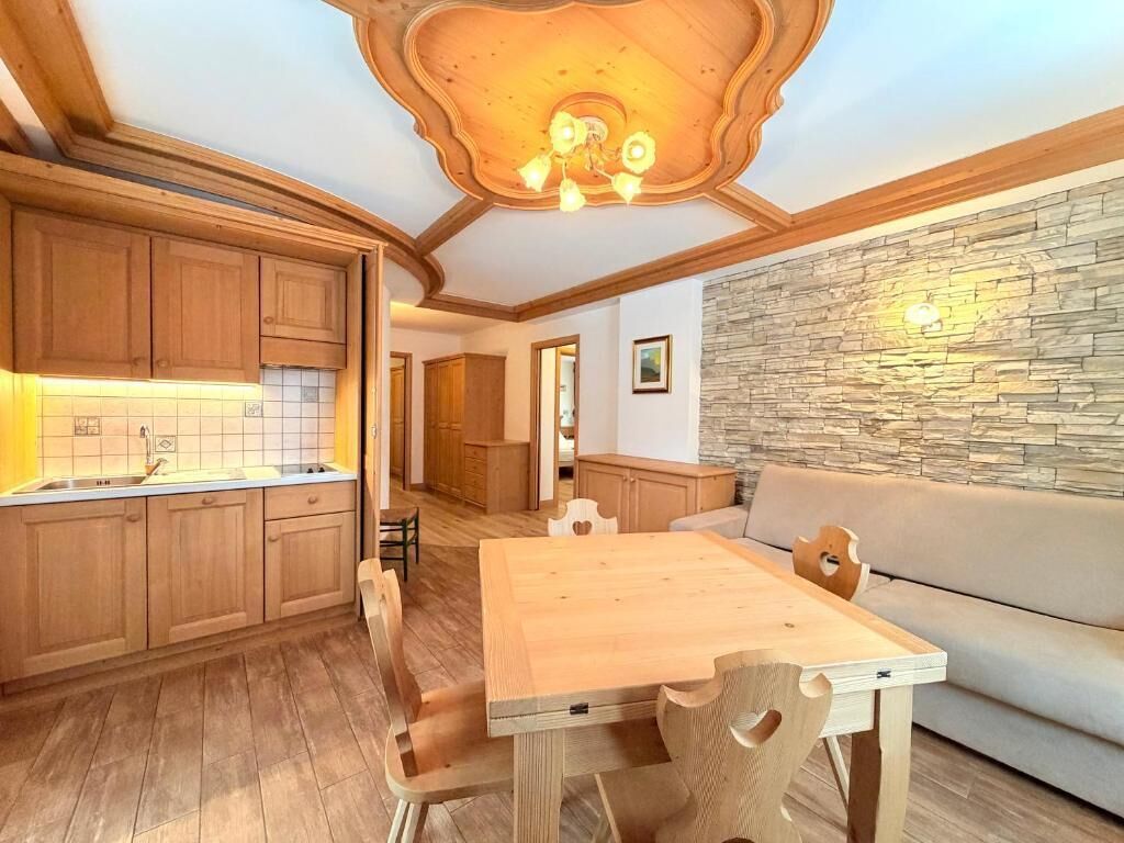A wooden dining table with chairs, a kitchen area with wooden cabinets, and a stone wall.