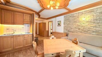 A wooden dining table with chairs, a kitchen area with wooden cabinets, and a stone wall.