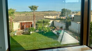A view through a large window of a residential area with a well-maintained lawn, a palm tree, and a building with a tiled roof. - Chicos de Lastanosa 4 (Lastanosa)