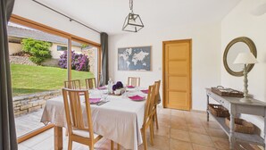 A dining area with a table set for four, a wooden chair, a lamp, and a view of the garden. - House for 8, fenced garden, terrace & BBQ (Fouesnant)