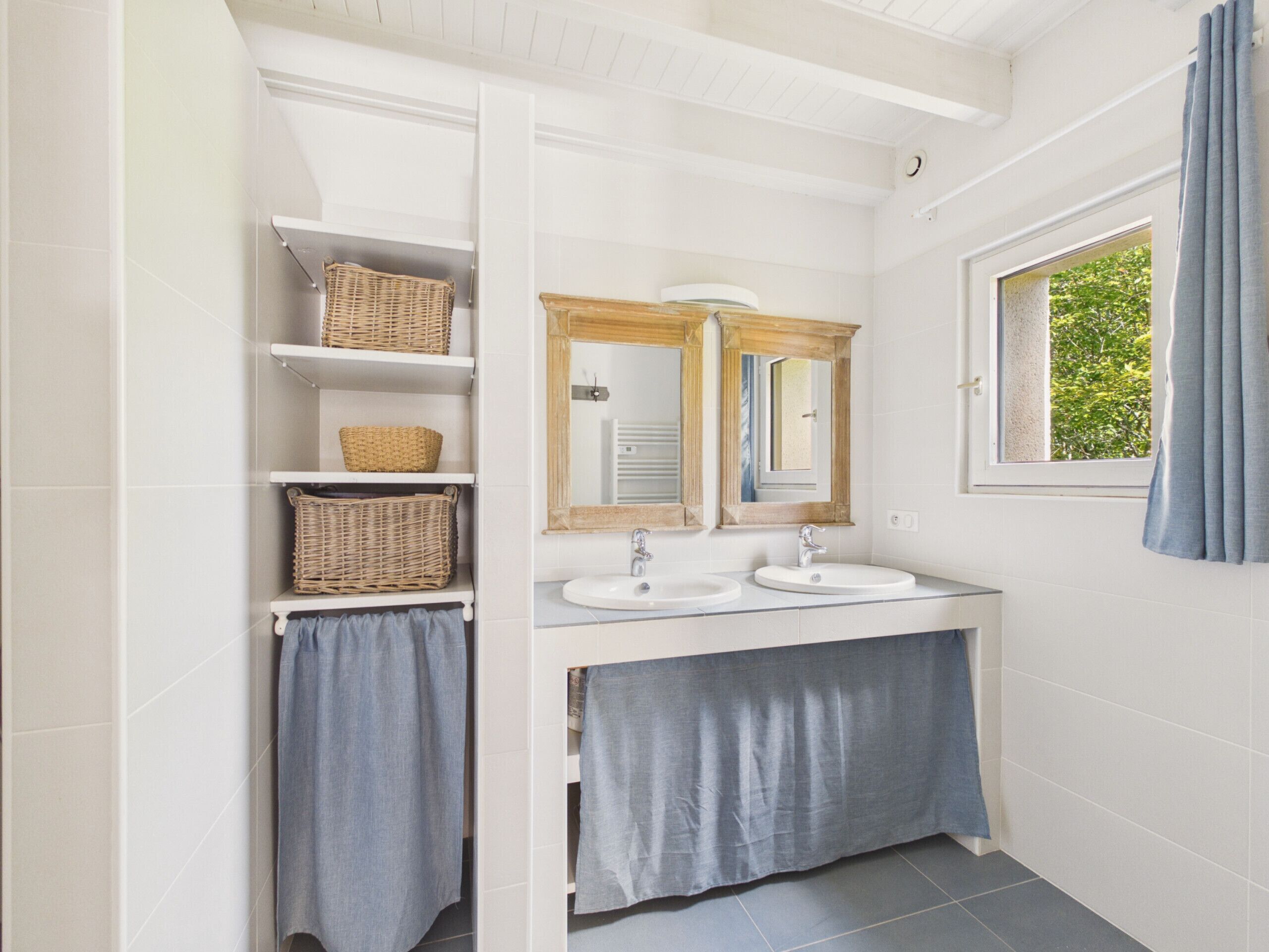 A bathroom with two sinks, a mirror, shelves with baskets, and a window with curtains.