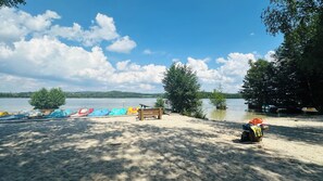 A lakeside beach with a row of colorful boats, a wooden bench, and a sandy area. - Brückelsee Chalet – Modern Holiday Home at Lake Brückelsee / Bavaria (Wackersdorf)