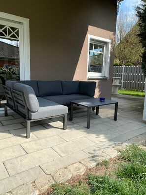 An outdoor patio with a gray sofa and a small table, a window, and a white fence in the background. - Brückelsee Chalet – Modern Holiday Home at Lake Brückelsee / Bavaria (Wackersdorf)