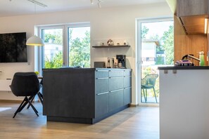 A modern kitchen with a central island, a dining area with a black chair, and a large window providing a view of the outdoors. - Murner See Lodge – Modern Holiday Apartment at the Lake "Murner See" / Bavaria (Wackersdorf)