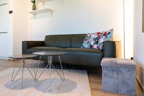 Ein modernes Wohnzimmer mit einem dunklen Sofa, zwei kleinen runden Tischen, einem grauen Hocker und einem weißen Teppich.