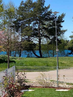 Ein von einer Hecke umgebener Parkbereich mit einer Lampe, Bäumen und einem Gewässer im Hintergrund.