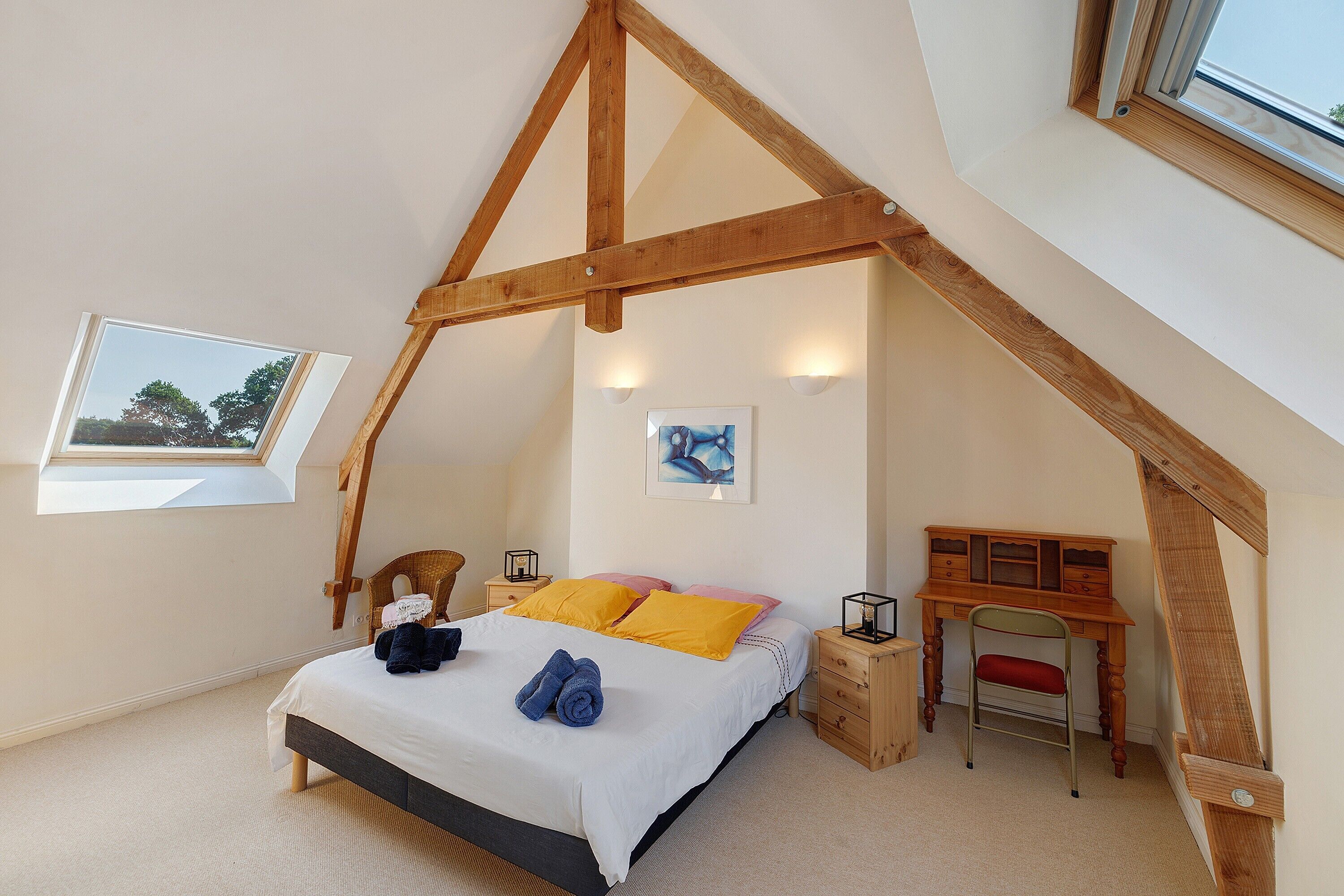 Une chambre avec un plafond en pente, un lit avec des oreillers, une chaise, un bureau et deux lampes fixées au mur.