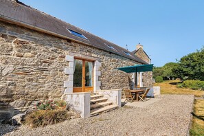 A stone building with a wooden door, a window with curtains, a table with chairs, and a green umbrella.