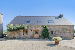 A stone building with a sloped roof, two windows, a wooden door, and a small garden area with gravel and a potted plant.