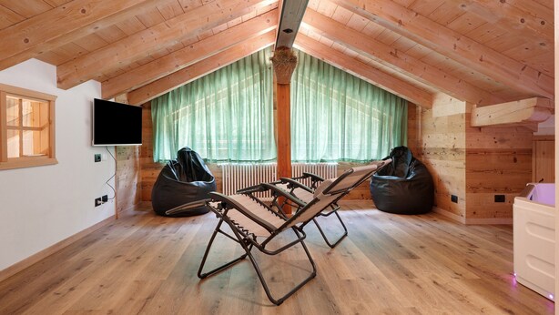 Una habitación con techo inclinado, vigas de madera, un ventanal con cortinas verdes, dos sillones de mimbre y un televisor de pantalla plana montado en la pared.