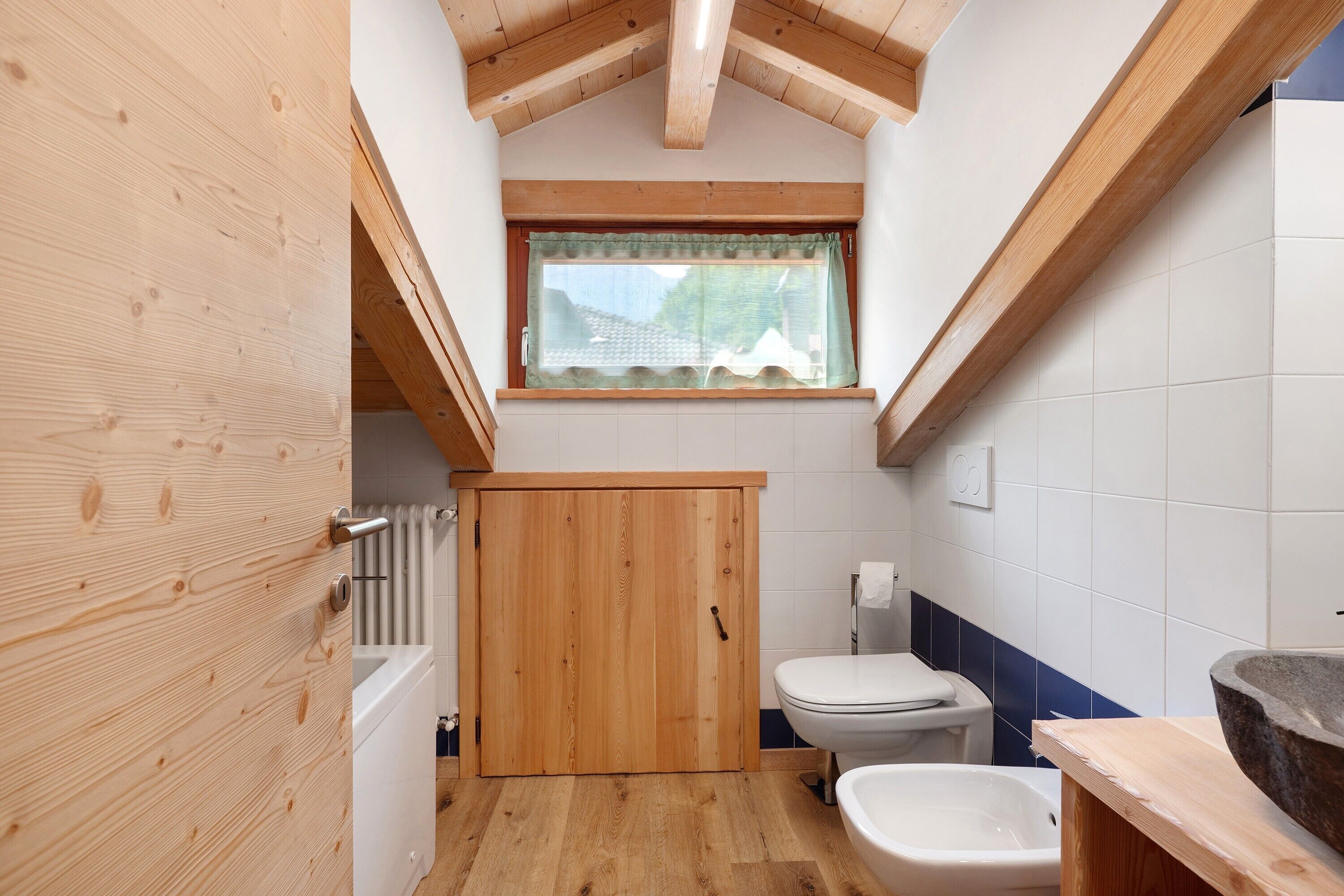 Ein kleines Badezimmer mit Holztür, einem Fenster mit Vorhang, einer Toilette und einem Bidet.