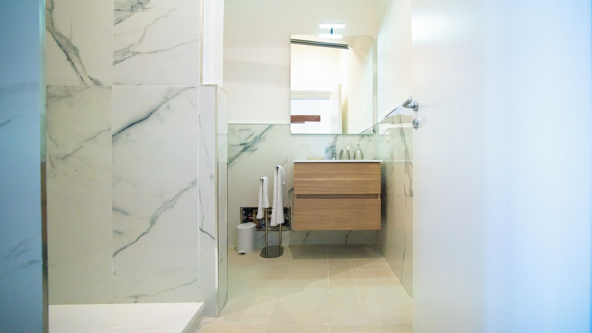 A bathroom with marble walls, a wooden vanity, a glass shower enclosure, and a mirror.