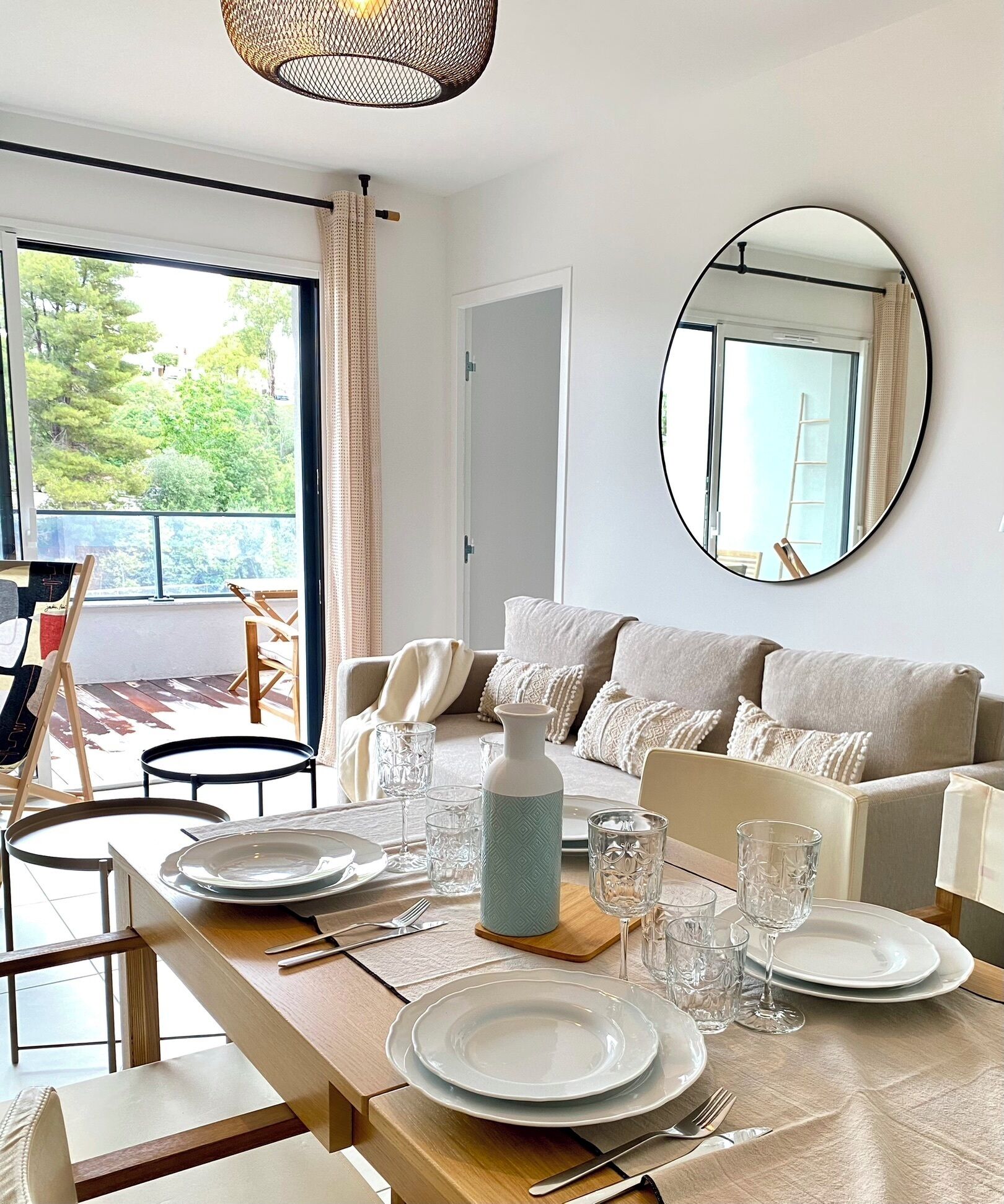 Un coin repas avec une table dressée pour quatre personnes, un canapé, un miroir et une vue sur la verdure à l’extérieur.