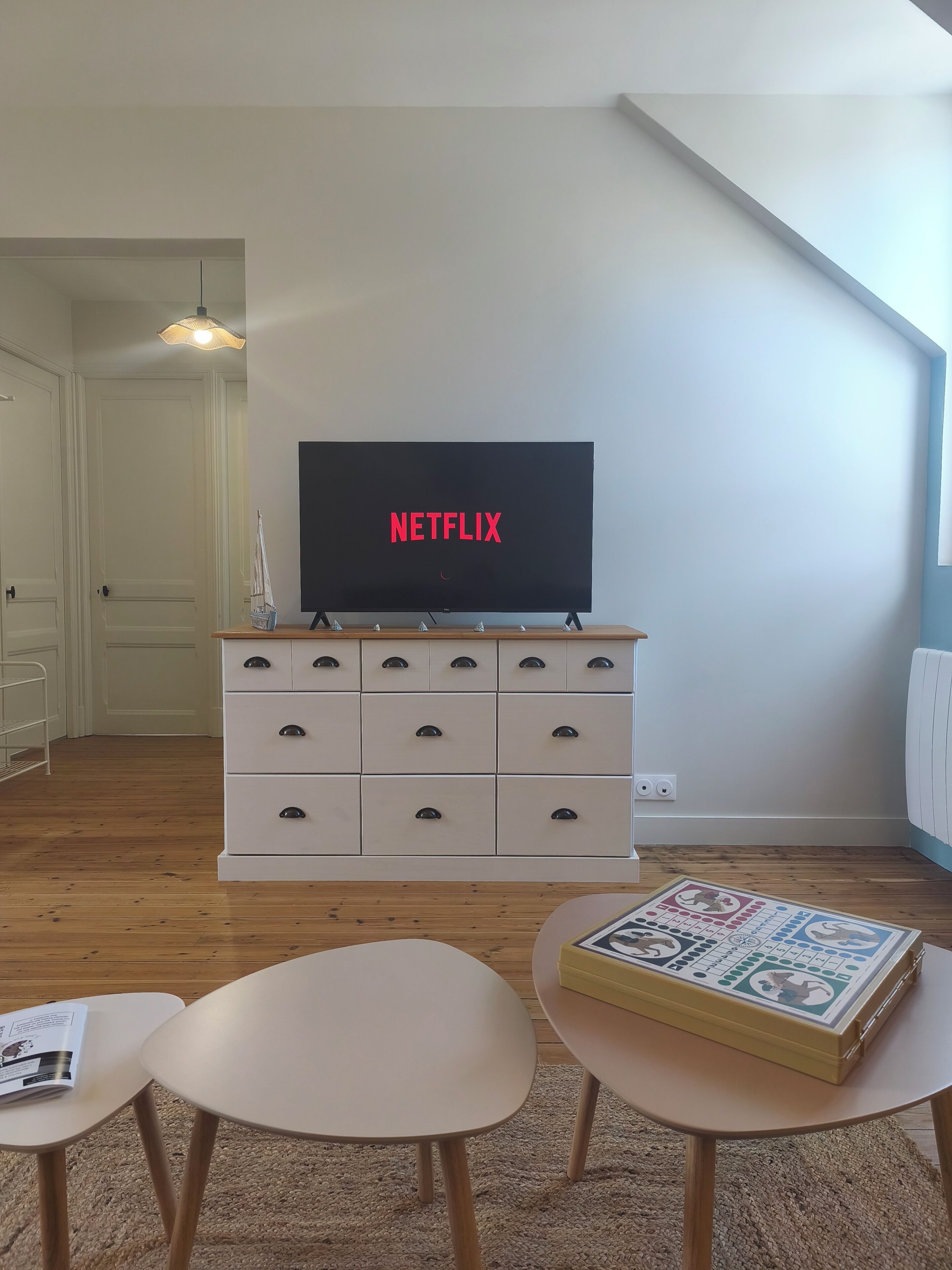 Uma sala de estar com uma TV exibindo 'NETFLIX', um aparador branco com gavetas, duas mesas redondas e um jogo de tabuleiro sobre uma delas.