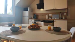 Et spisebord med servietringe, skåle og glas med appelsinjuice, et køkken med hårde hvidevarer, samt et vindue med dagslys.
