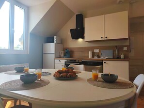 A dining table with placemats, bowls, and glasses of orange juice, a kitchen area with appliances, and a window with daylight. - Écume and Horizon—3-bedroom apartment for up to 4 people—Beach 100 meters away—Ocean view from the living room (Ault)