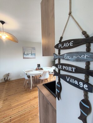 A dining area with a white table and chairs, a wooden floor, and a wall-mounted sign with words 'PLAGE la mer,' 'PORT,' and 'Village.' - Écume and Horizon—3-bedroom apartment for up to 4 people—Beach 100 meters away—Ocean view from the living room (Ault)