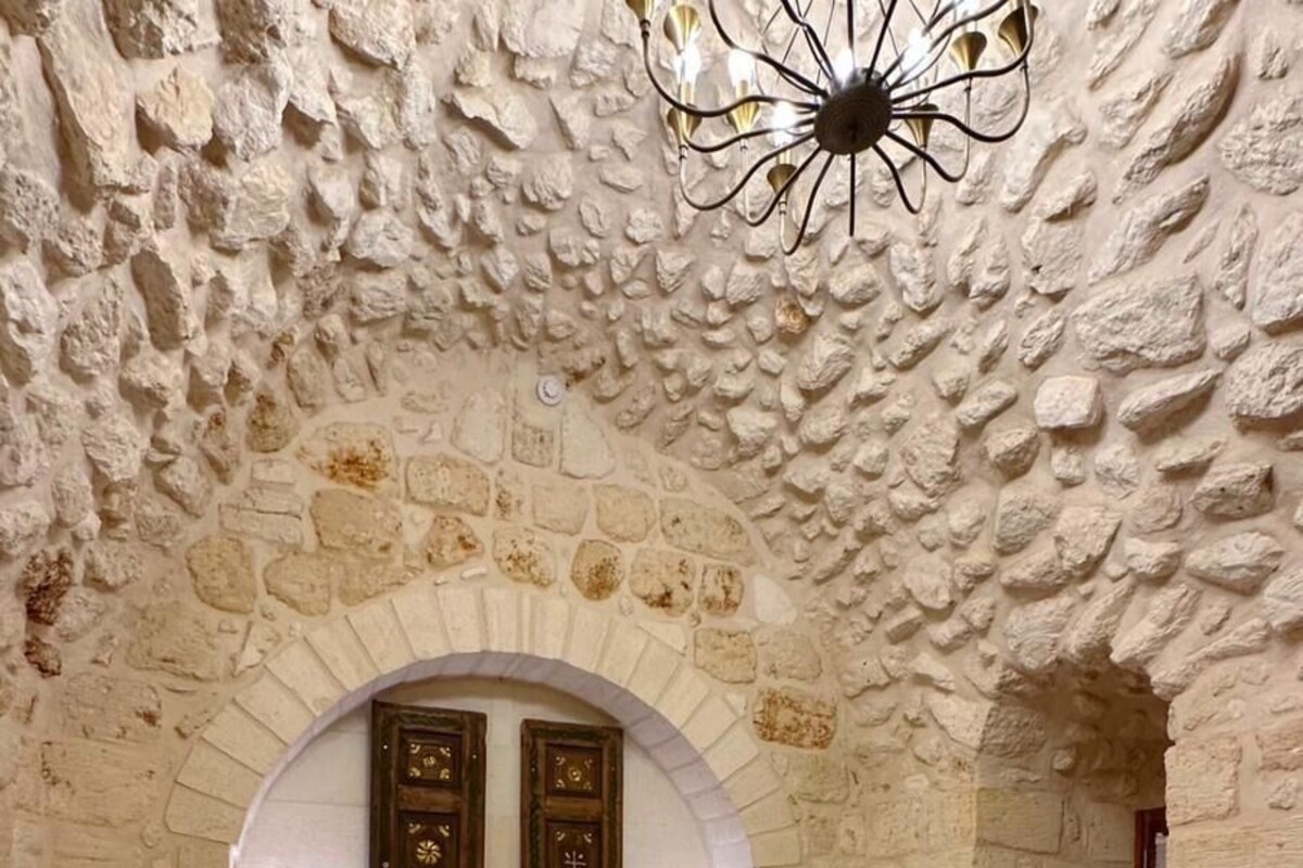 Quarto com parede de pedra, porta em arco, cama com roupa de cama branca e um candelabro.