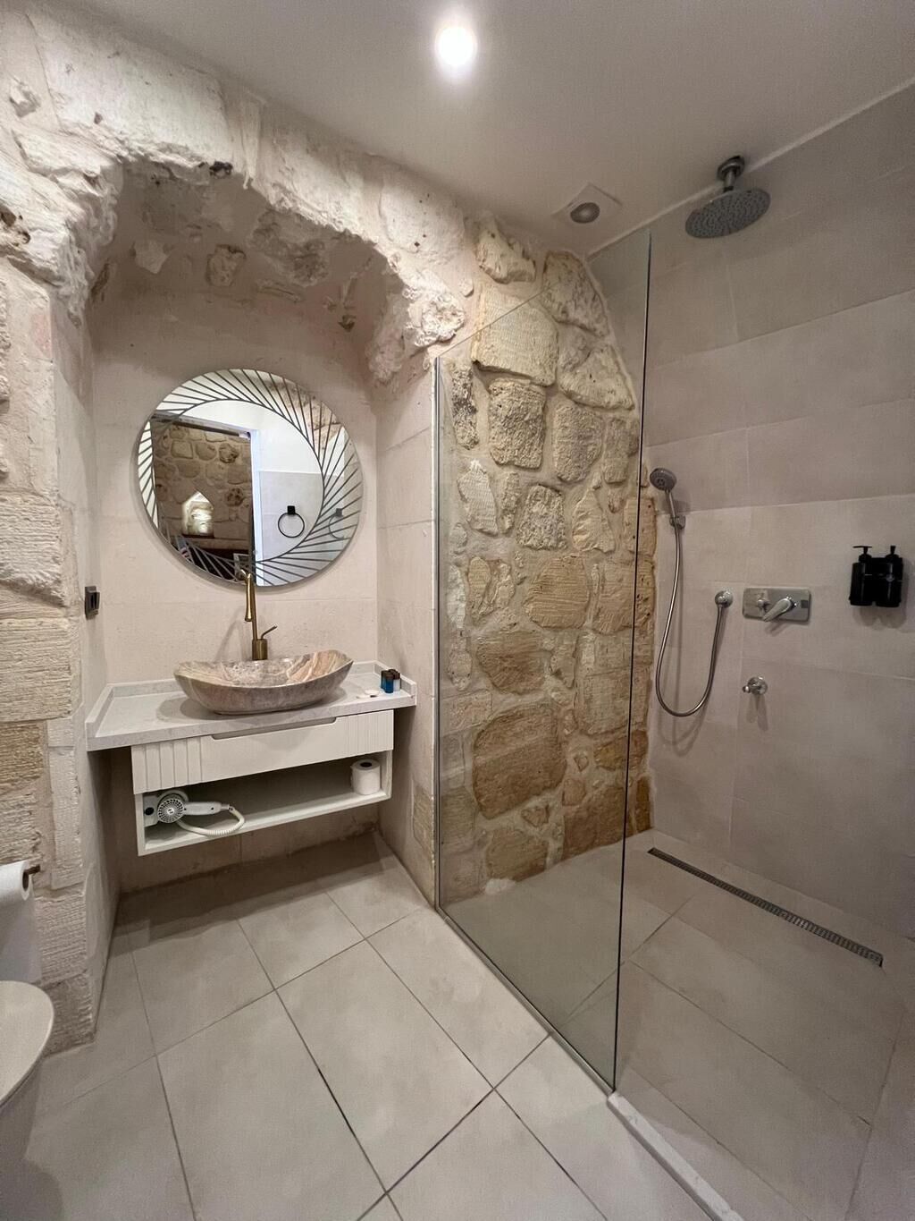 Un baño con pared de piedra, un espejo redondo, un lavabo con cuenca y una ducha.
