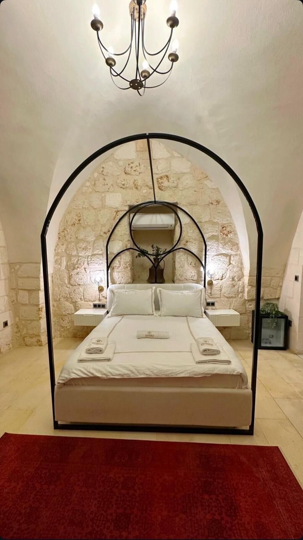Een slaapkamer met een stenen muur, een kroonluchter, een bed met witte lakens en een rood tapijt.