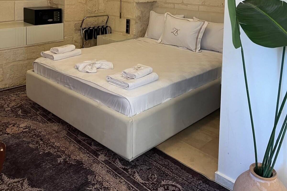 Quarto com parede de pedra, cama com roupa de cama branca, mesa de cabeceira e uma planta grande.