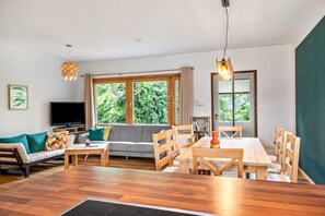 A living room with a sofa, TV, wooden dining table, chairs, and large windows. - Gina's Holiday Home with Garden, Treehouse Terrace and Sauna (Eggstedt)