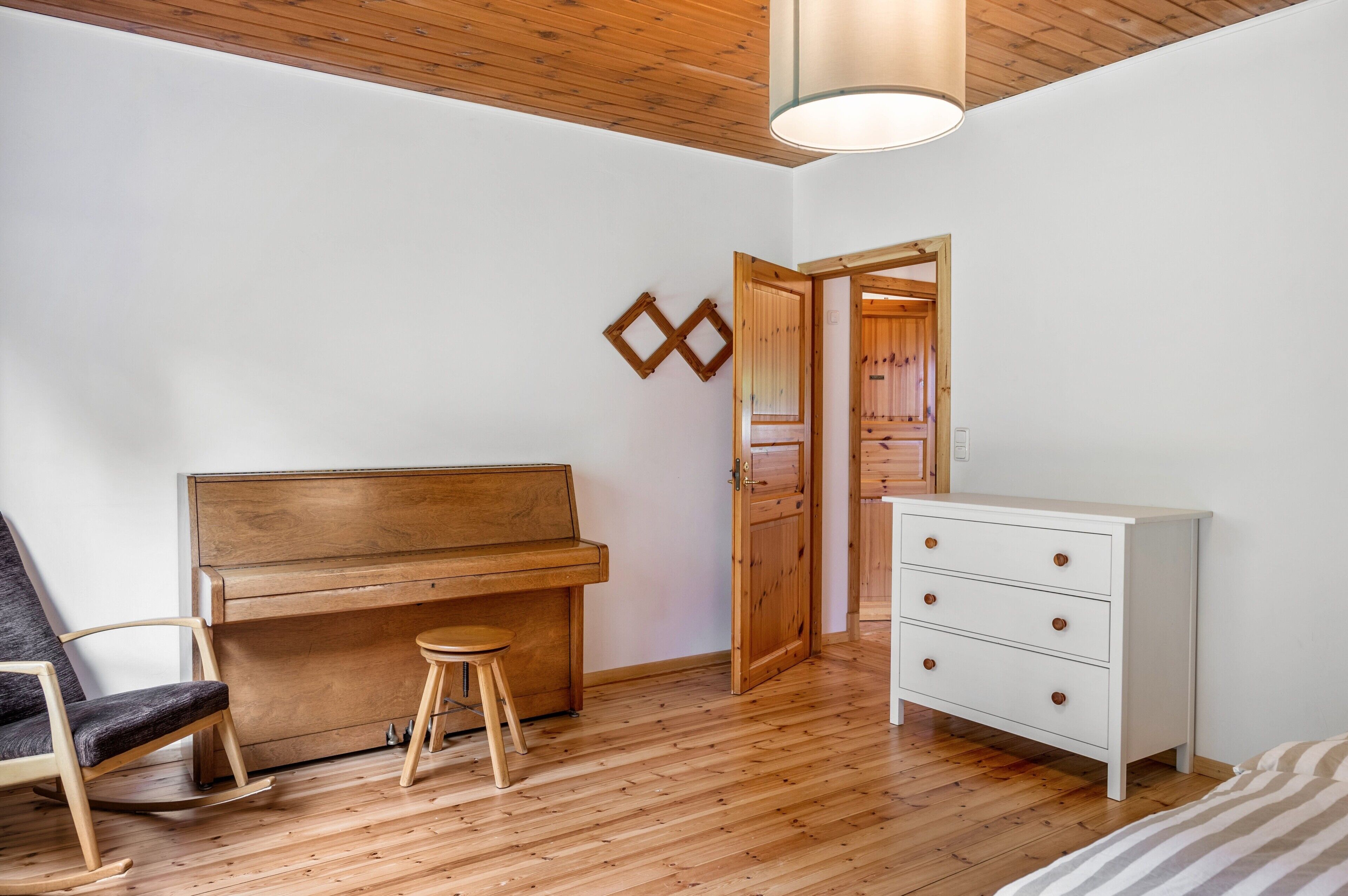 Een kamer met een houten piano, een witte ladekast, een schommelstoel en een houten kruk.
