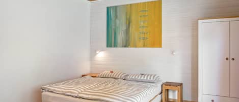 Un dormitorio con una cama, un taburete de madera, un armario y un cuadro en la pared.
