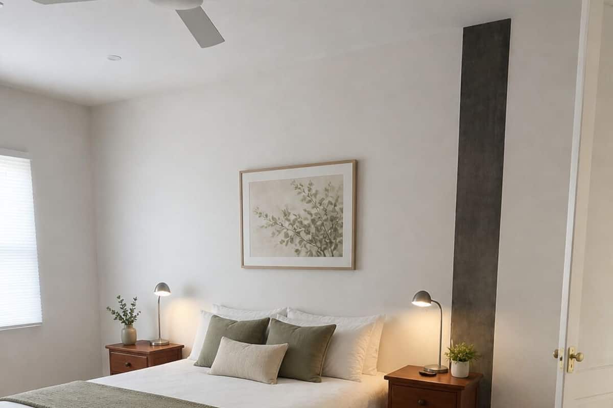Un dormitorio con cama, mesitas de noche, ventilador de techo, un cuadro enmarcado y una puerta.