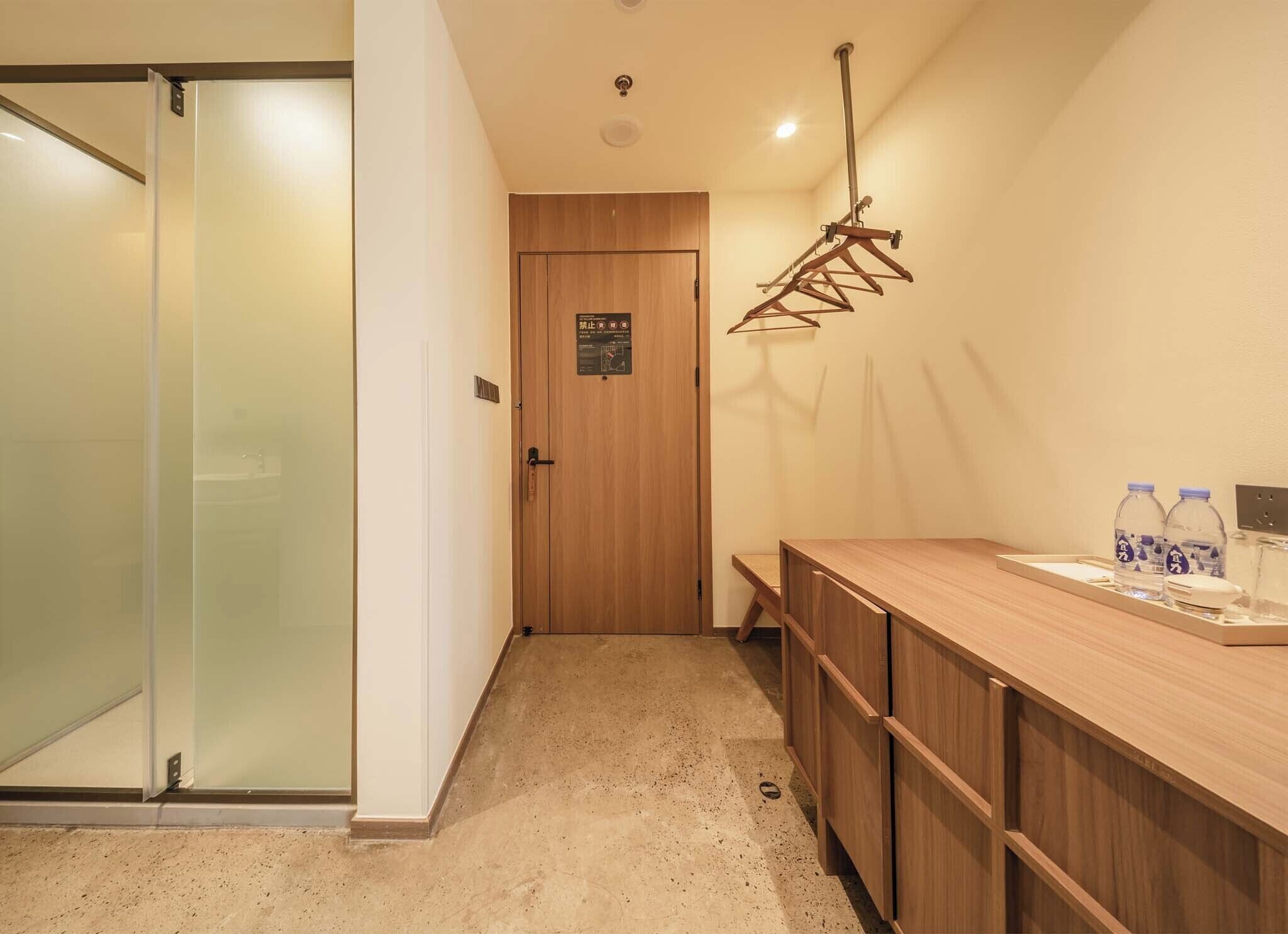 A hotel room with a wooden door, a coat rack, a wooden counter with a tray, and a water bottle.