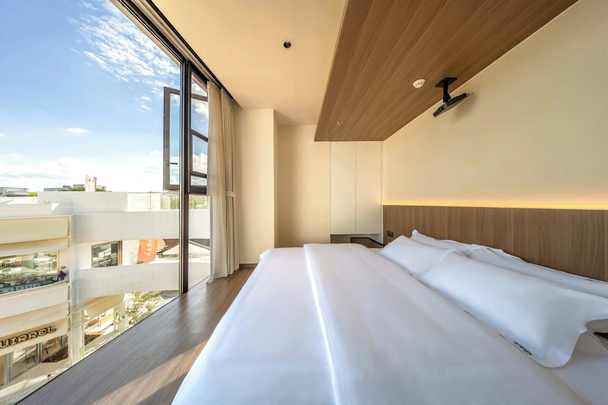 A modern hotel room with a large bed, a wooden headboard, a nightstand, and a view of the city through large windows.