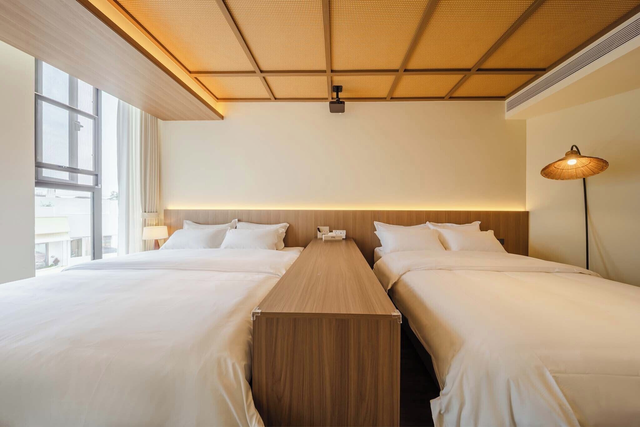 Une chambre d’hôtel avec deux lits, une tête de lit en bois, une longue table en bois, une lampe et une grande fenêtre.