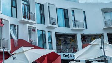 Un edificio moderno con varios pisos, ventanas grandes y mobiliario exterior con sombrillas.