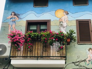 Un edificio con un mural, un balcón de madera con flores rosadas y una ventana con contraventanas marrones.