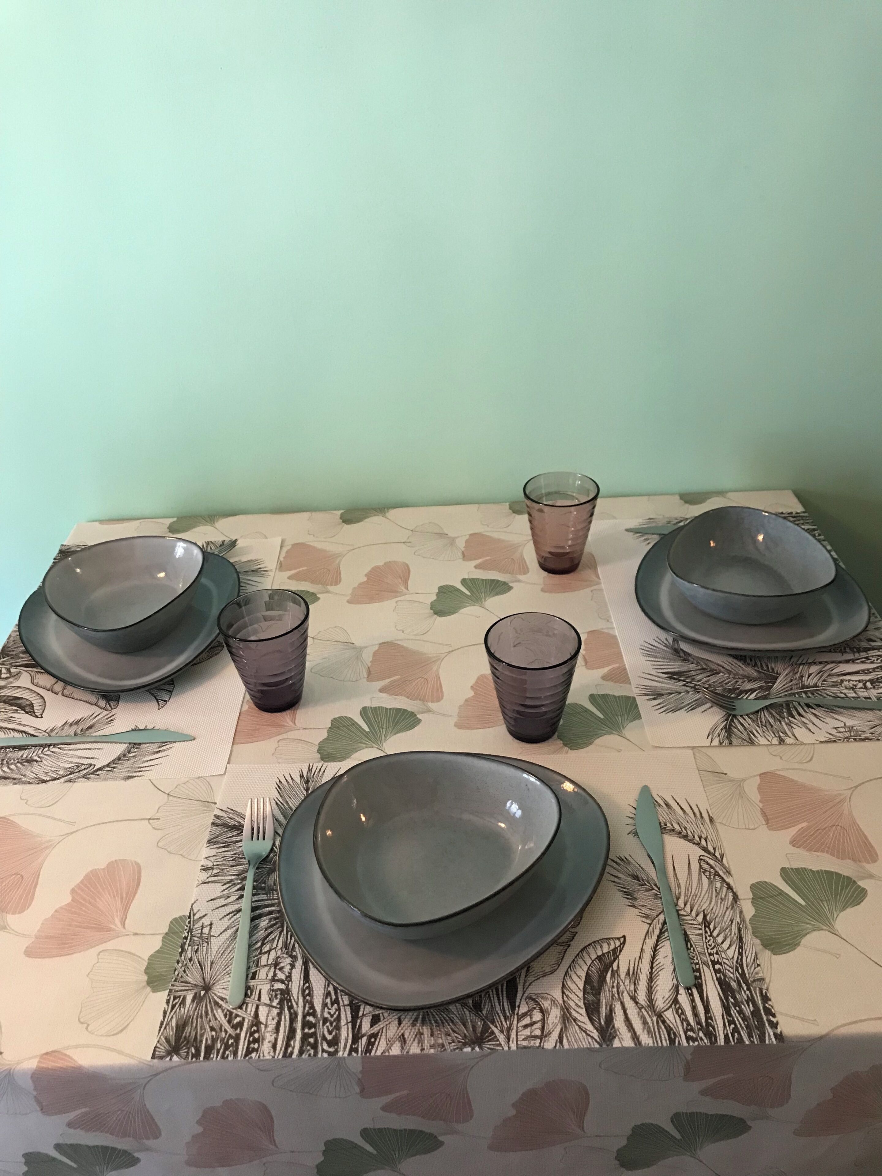 A dining table with plates, glasses, and cutlery set on a tablecloth with a leaf pattern.