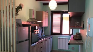 A kitchen with a refrigerator, wooden cabinets, a window with blinds, a hanging light fixture, and a radiator.