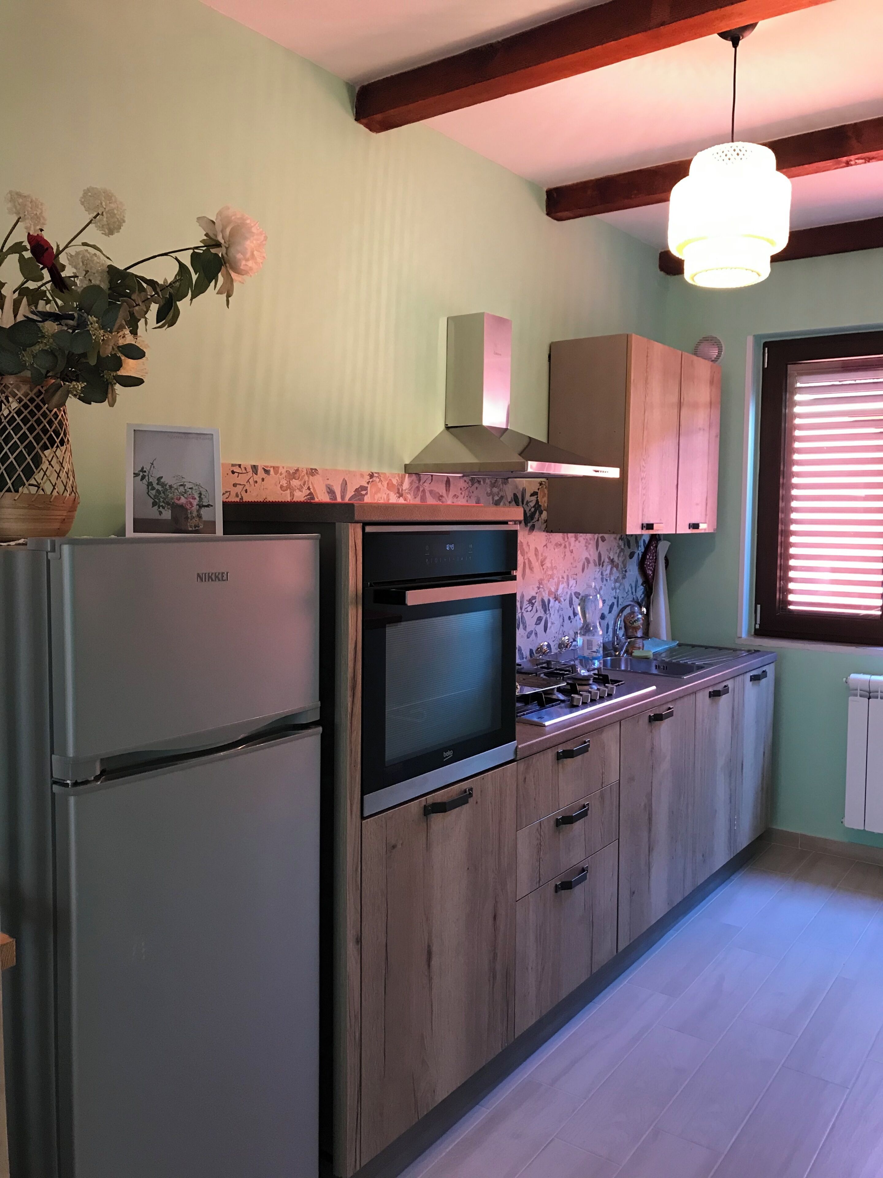 Eine Küche mit Einbauofen, Kühlschrank, Holzschränken, einer Hängeleuchte und einem Fenster mit Jalousien.