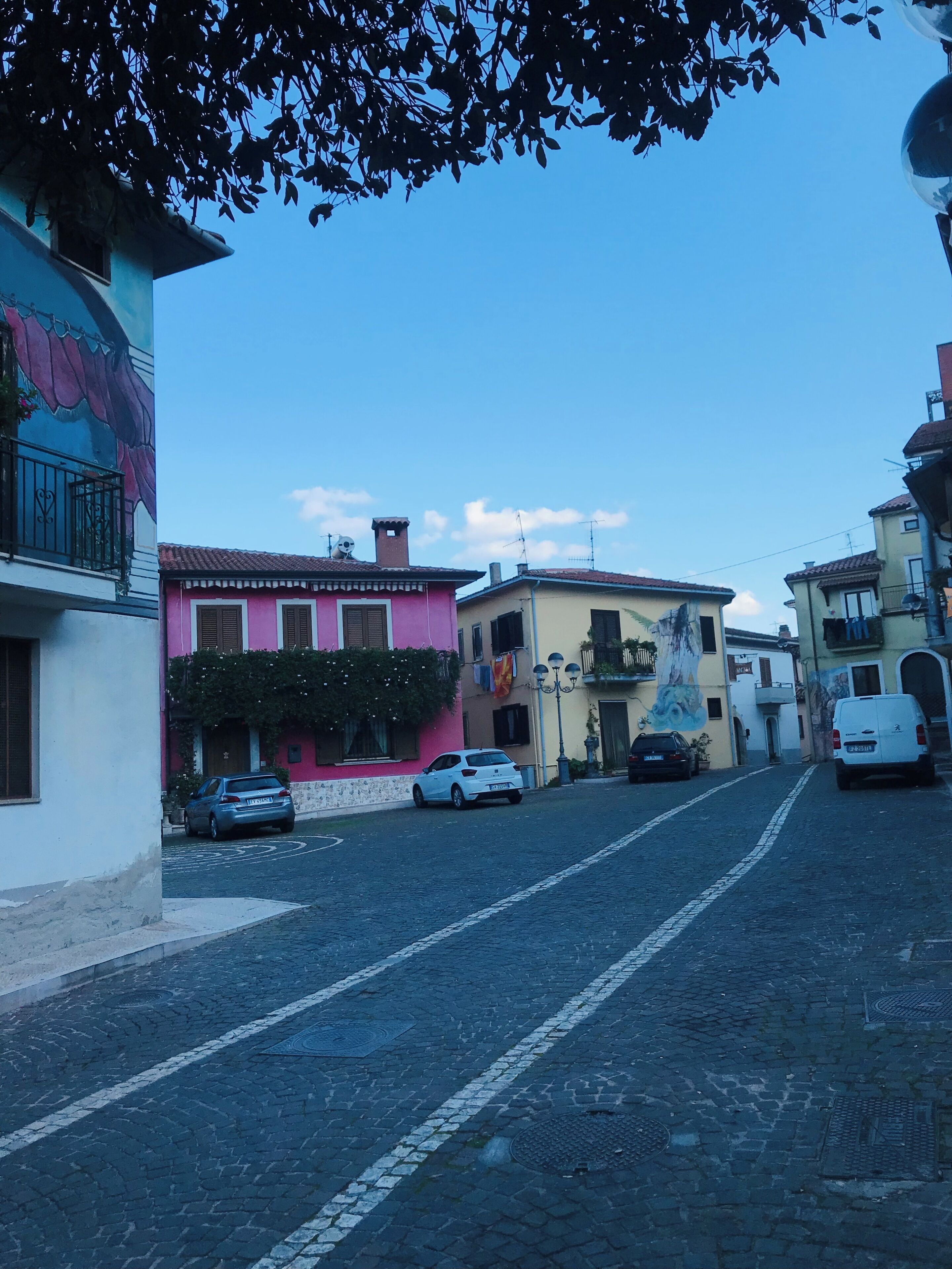 A cobblestone street lined with colorful buildings, parked cars, and a clear blue sky.
