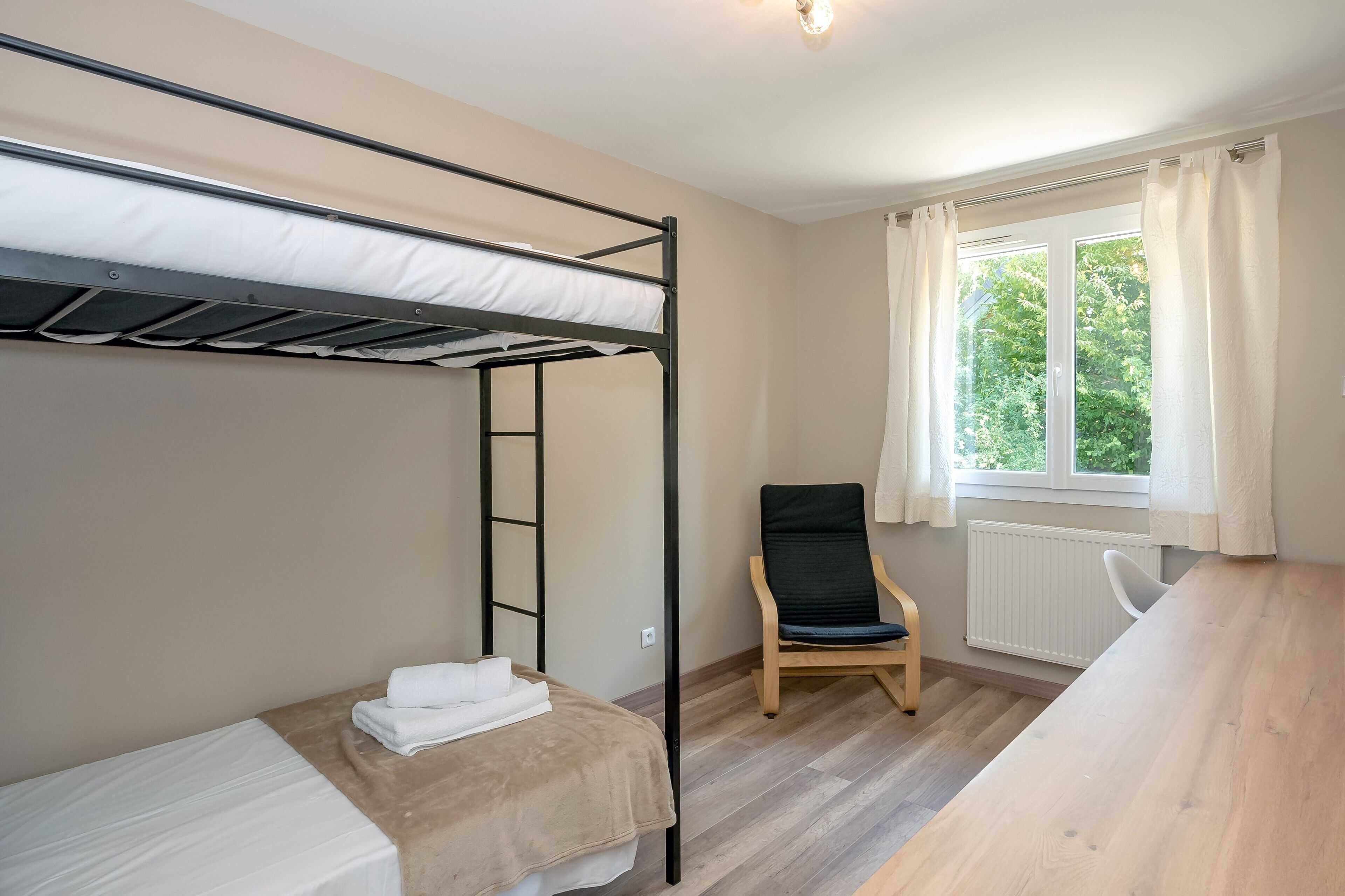 A bunk bed room with a wooden desk and a chair, a window with curtains, and a radiator.