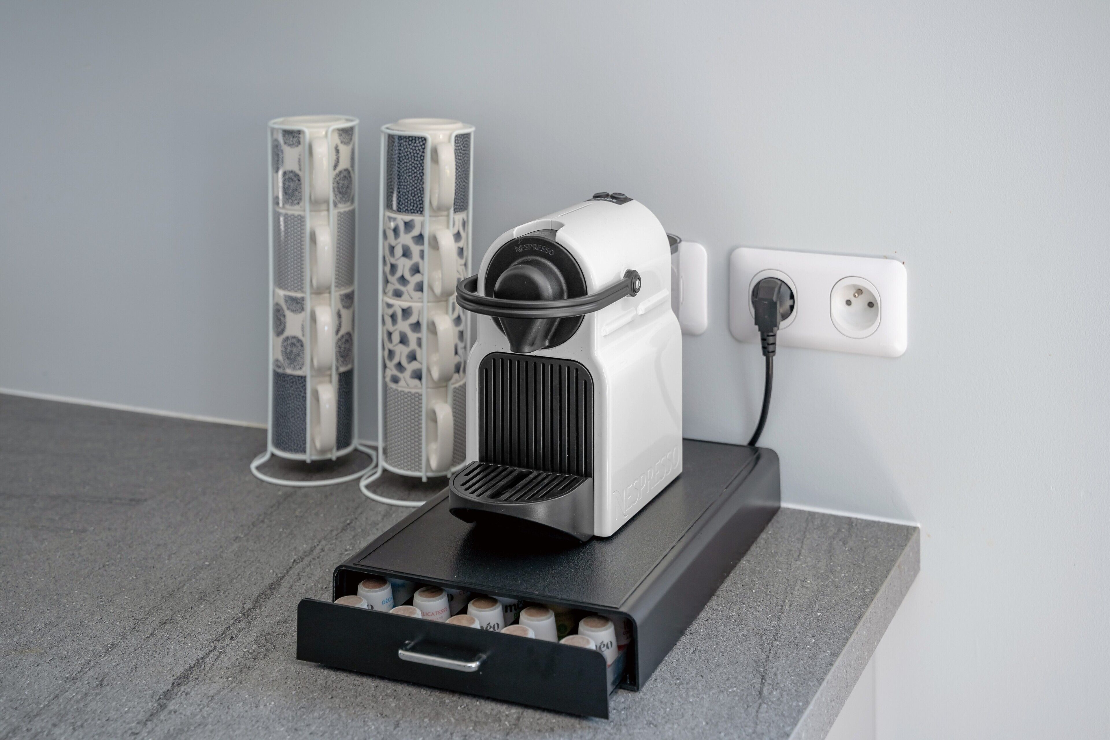 A coffee machine with a drawer for coffee pods, connected to a power outlet, and two cylindrical containers with a speckled pattern.