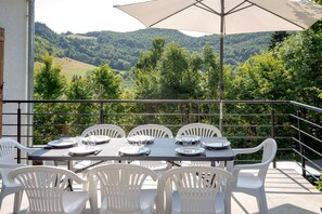 Une terrasse avec une table dressée pour quatre personnes, entourée de chaises blanches, donnant sur une vallée verdoyante.